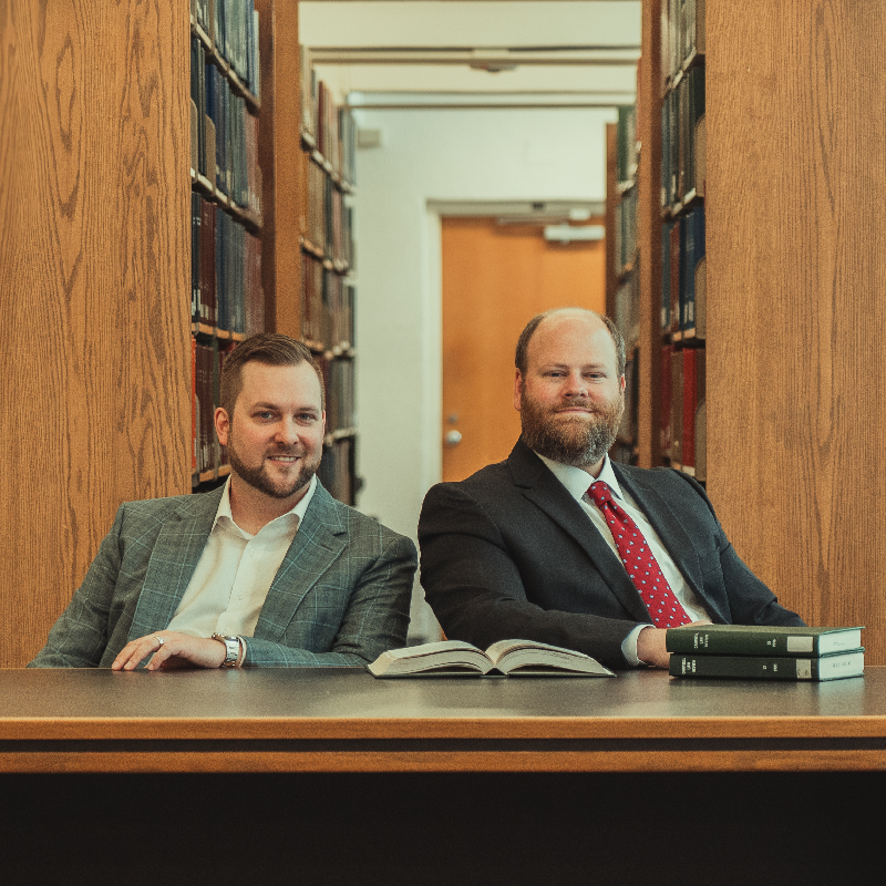 JJ & Harrison in suits seated at a desk in a law library, surrounded by law books