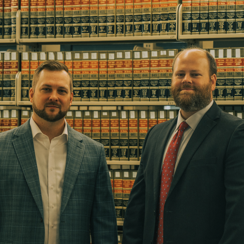 JJ & Harrison in suits stand professionally in front of a law library bookshelf filled with legal books and resources.