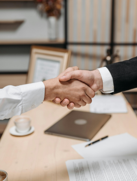 Two people in business attire shaking hands over a desk with a laptop, documents, pen, and a cup of coffee, suggesting a successful meeting or agreement.